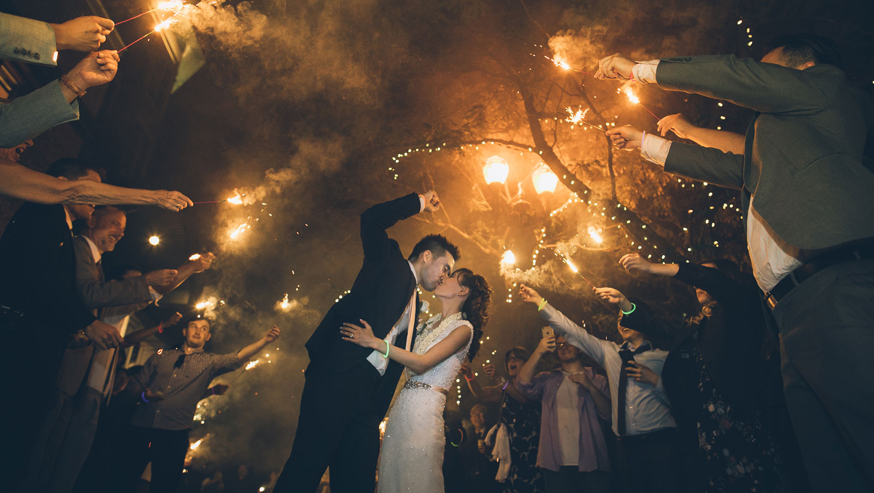 newlywed couple kissing while guests hold lit sparklers over their heads outside at night