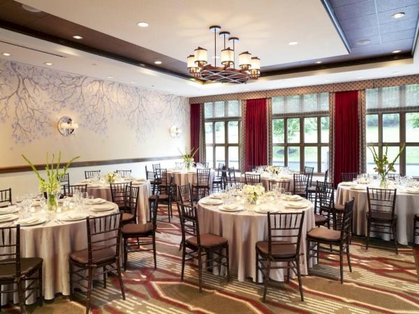 A stylish banquet hall with round tables, black chairs, white tablecloths, a chandelier, and tall windows with red curtains.
