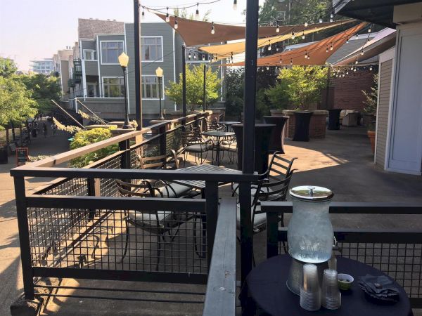 A sunny outdoor patio with string lights, tables and chairs, and an urban brick courtyard surrounded by trees.