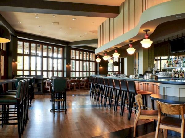 A modern hotel bar interior with a long counter, stools, pendant lights, and large windows along the wall.