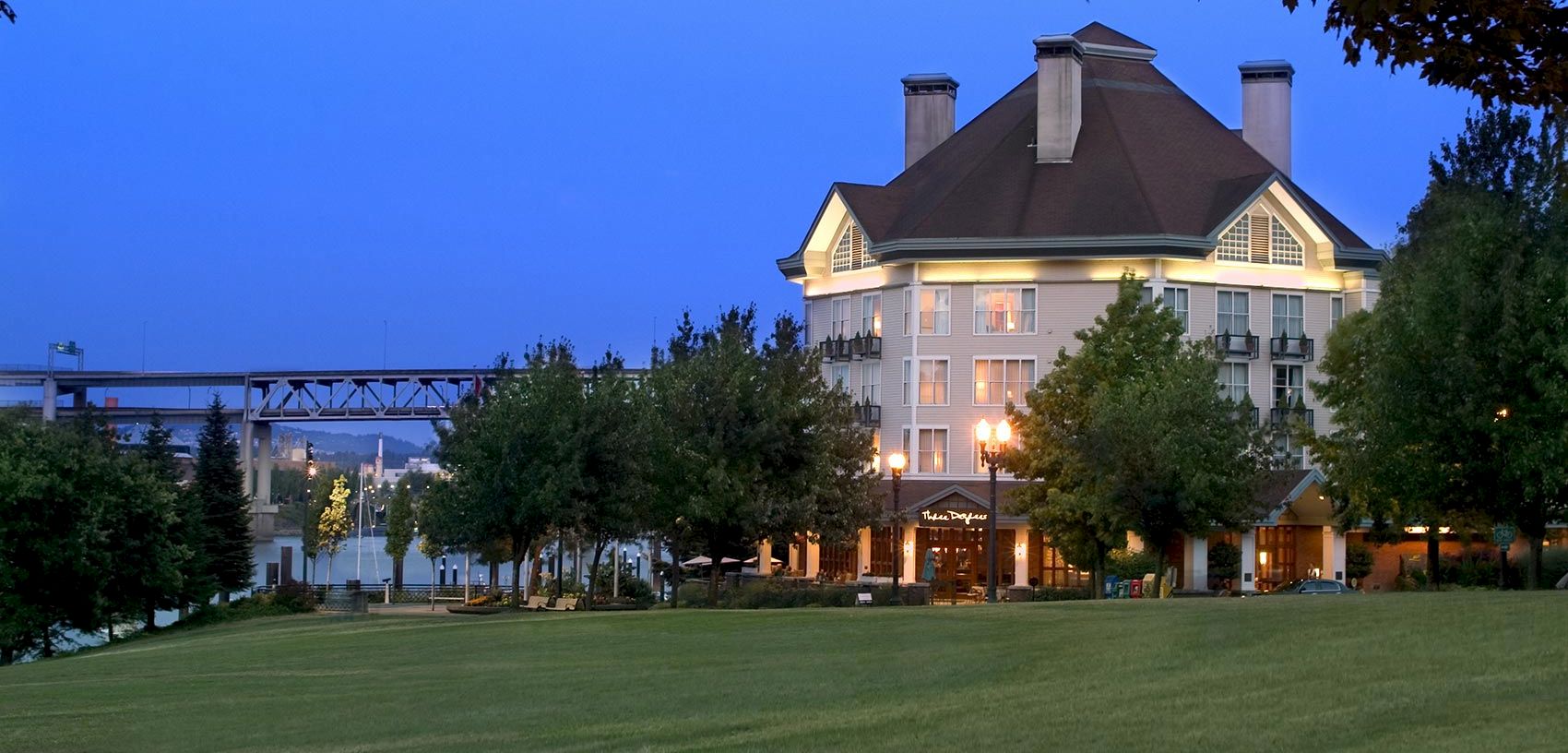 A riverside hotel at dusk with bright lamps, surrounding trees, and a manicured lawn under a clear blue sky.