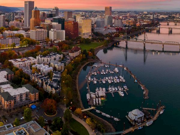 An aerial view at dusk of a riverside city skyline, bridges, and a marina packed with docked boats along the waterfront.