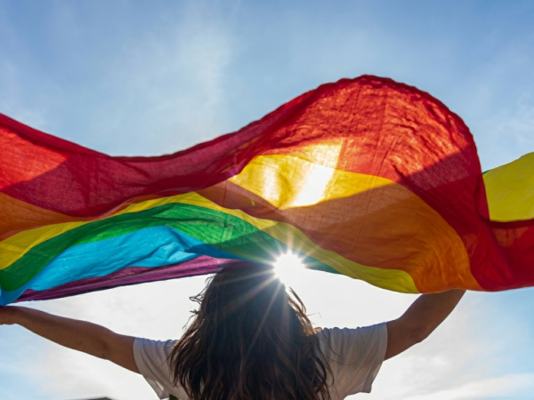 A person holds a bright rainbow fabric kite/parachute above their head under a blue sky.