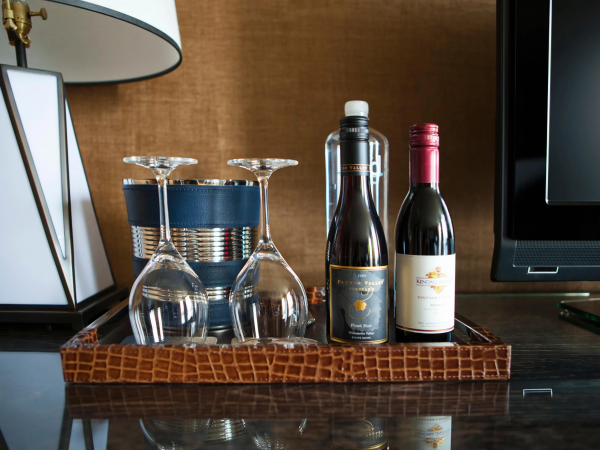 Two wine bottles and four empty stemmed glasses sit on a tray beside a lamp and a TV in a cozy room.