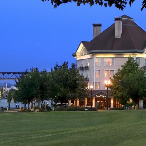 A large hotel-style building with lit windows at dusk, surrounded by trees and a grassy lawn.