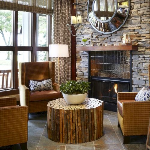 A cozy living room with a stone fireplace, armchairs, patterned lamps, and a wooden-log coffee table by large windows.