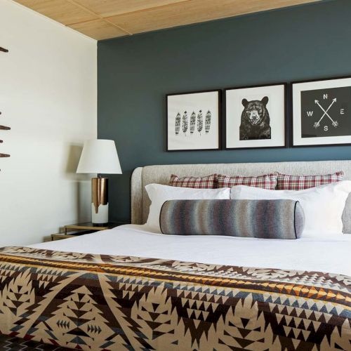 A cozy modern bedroom with a neatly made bed, two lamps, framed animal and compass wall art, and window blinds.