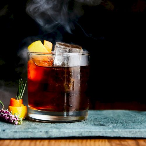 A smoked glass of red iced tea or cocktail with citrus slices, sitting on a wooden table with flowers nearby.
