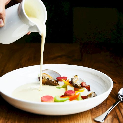 A hand pours creamy milk into a bowl of mixed seafood and vegetables on a wooden table.