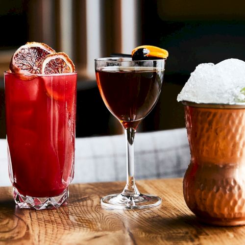 Three colorful cocktails on a wooden bar table&mdash;red, dark, and a copper drink with crushed ice and a lime garnish.
