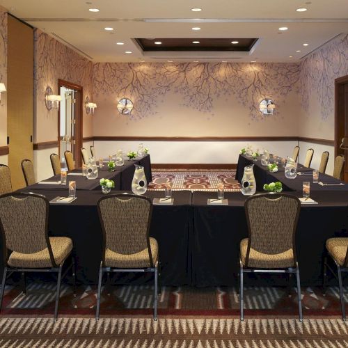 A formal meeting room with a long banquet table set with water bottles, flowers, chairs, and patterned wallpaper and lighting.