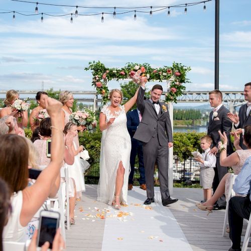 Outdoor wedding with bride and groom under a flower arch, guests clapping on a deck by the water.