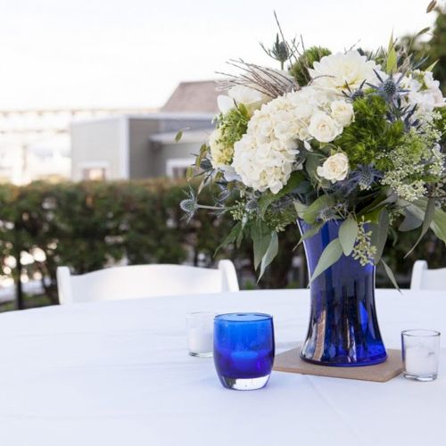A blue glass vase with a white floral bouquet sits on a decorated outdoor table with blue drinks and glassware.