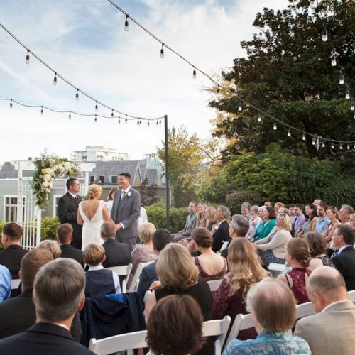 An outdoor wedding ceremony with a bride and groom at the front, guests seated, and string lights hanging overhead.