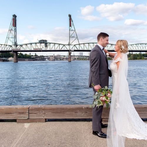 Bride and groom pose by a river with a steel bridge in the background, smiling and holding a bouquet.