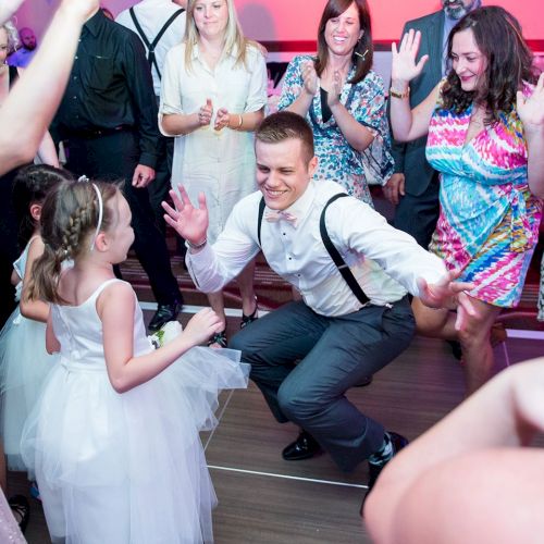 A lively wedding celebration: guests cheer and dance as a man kneels and a flower-girl in white bows around him.