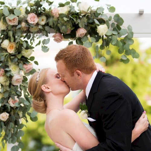 A bride and groom kiss beneath an elegant floral arch at an outdoor wedding ceremony.