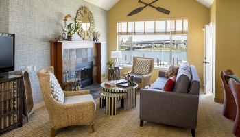 A cozy living room with a stone fireplace, wicker chairs, striped table, and large window showing a river view.