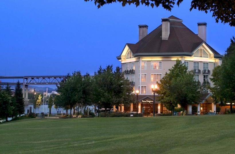 A large hotel building glows at dusk behind trees, with a bridge and river in the background and a green lawn in front.