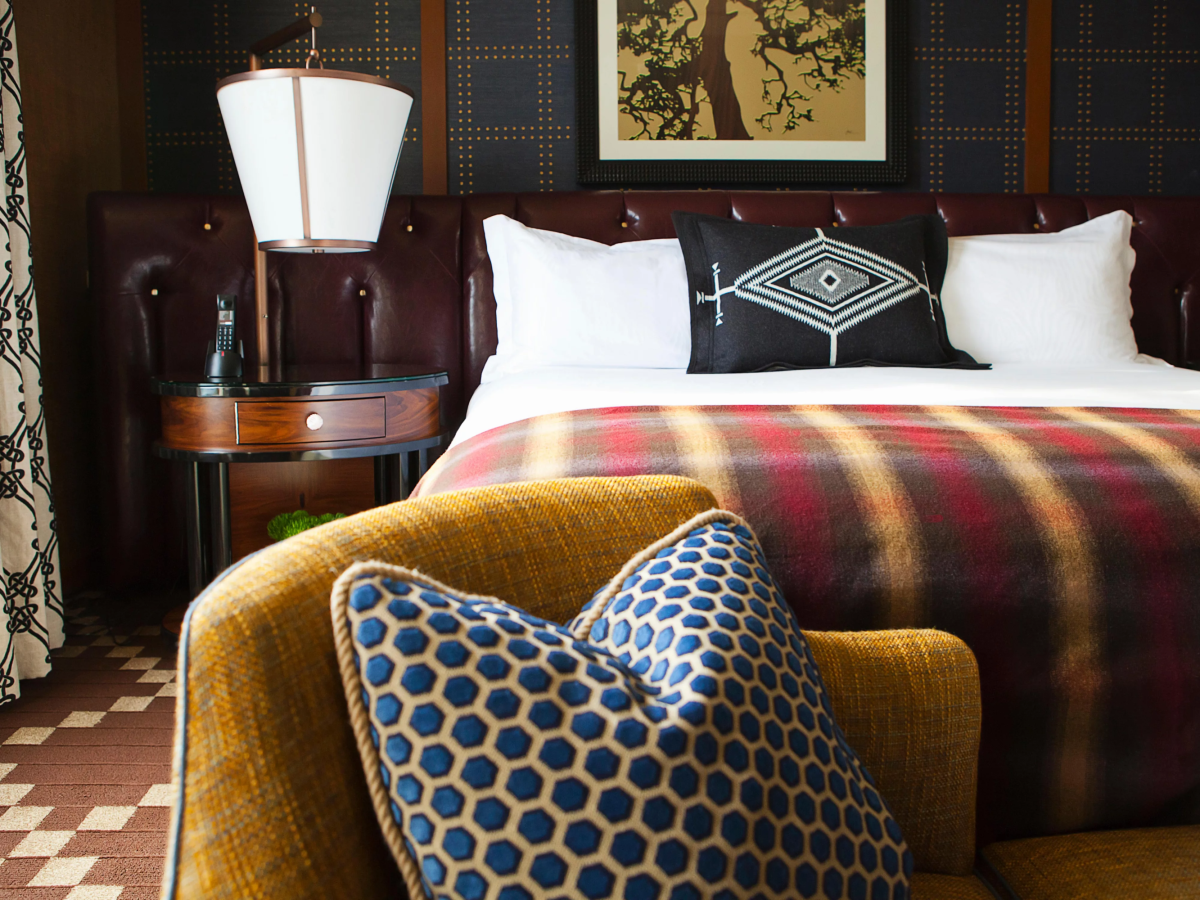 A cozy hotel bedroom with a leather tufted headboard, lit bedside lamp, framed artwork, and neatly made bed with patterned pillows.
