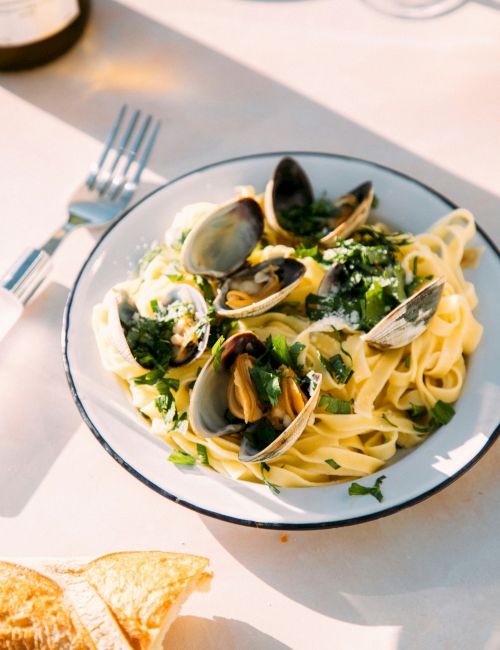 A bowl of fettuccine with clams and mussels, topped with parsley and lemon on a sunny table.
