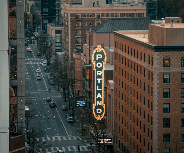 A busy city street at dusk beside a tall &ldquo;PORTLAND&rdquo; neon sign on a building, with cars and high-rise offices around it.