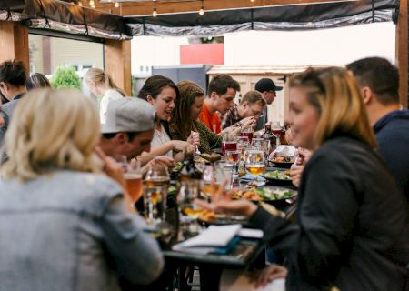 People gather at an outdoor patio table, eating and drinking craft beverages under string lights at a casual event.